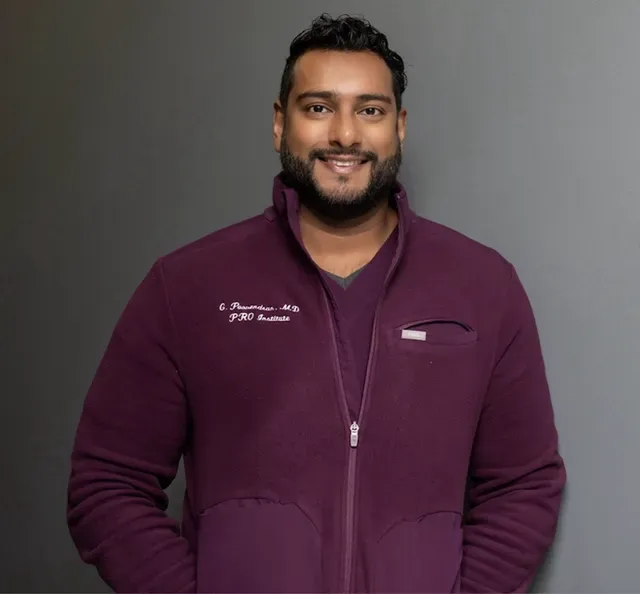 Smiling man wearing a maroon zip-up jacket with “C. Poovendran, M.D.” and “PRO Institute” embroidered on the chest, standing against a plain gray background – poovendran